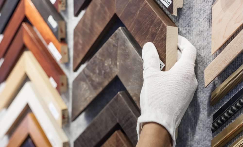 A person wearing a white glove picks up a sample wood frame piece from a wall of wood frame samples in various colors and treatments.