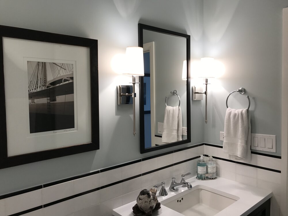 A clean, modern bathroom displays a mirror with a black frame that features long rod light sconces on either side of the mirror.
