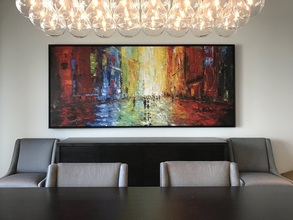 A meeting room with a horizontal colorful painting hung on the wall. Plus, there is a glass bulb chandelier, a wood table, and chairs.