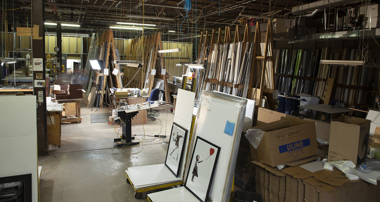 A workshop or storage room full of wood framing supplies, including wood frames, cardboard boxes, mounting and framing materials, and more.