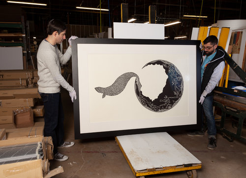 Two men hold up a large framed art piece in a black frame. The black and white art piece shows a seal-shaped animal leaving a moon shape.
