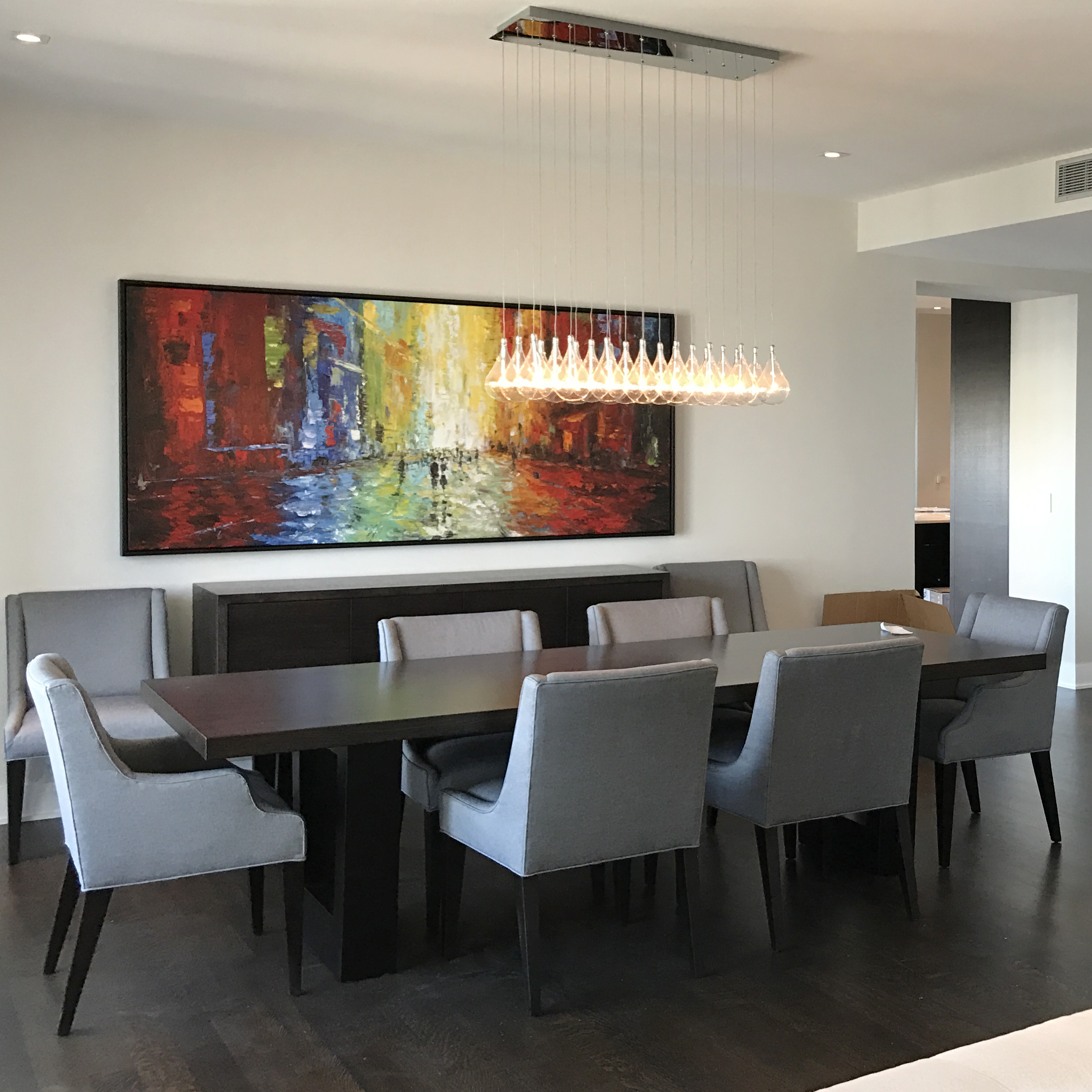 A meeting room has a long rectangular table with several gray chairs in the middle. A large, horizontal, colorful art piece is on the wall.