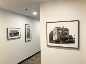 A plain office building hallway features black and white framed photographs hanging on the walls. Photos show places in the past.