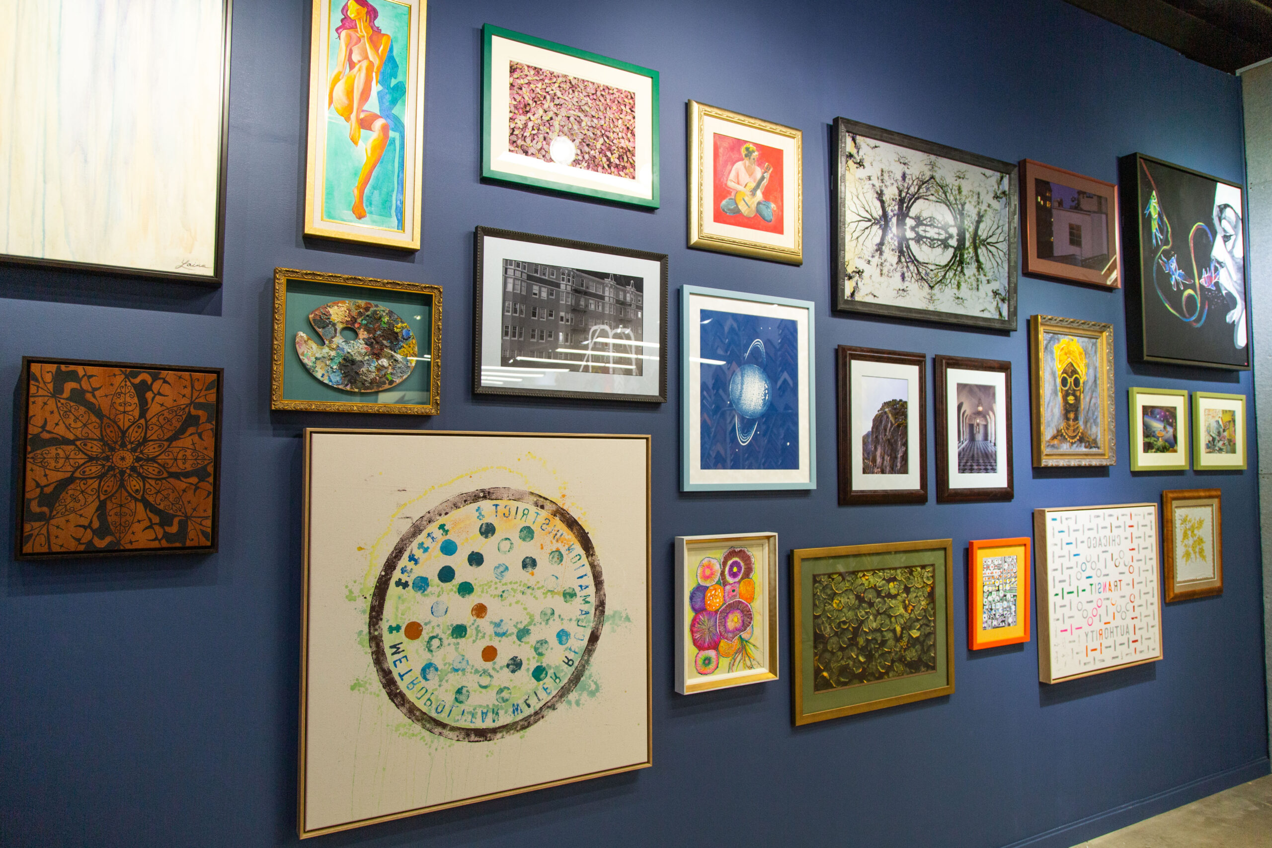 A blue-painted wall features a variety of framed art pieces. The collection of art pieces is all different and has different colored frames.