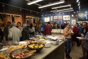 People gather in the Peterson Picture Co. in-house art gallery, looking at art framed on the blue pained walls and eat from a buffet table full of food.
