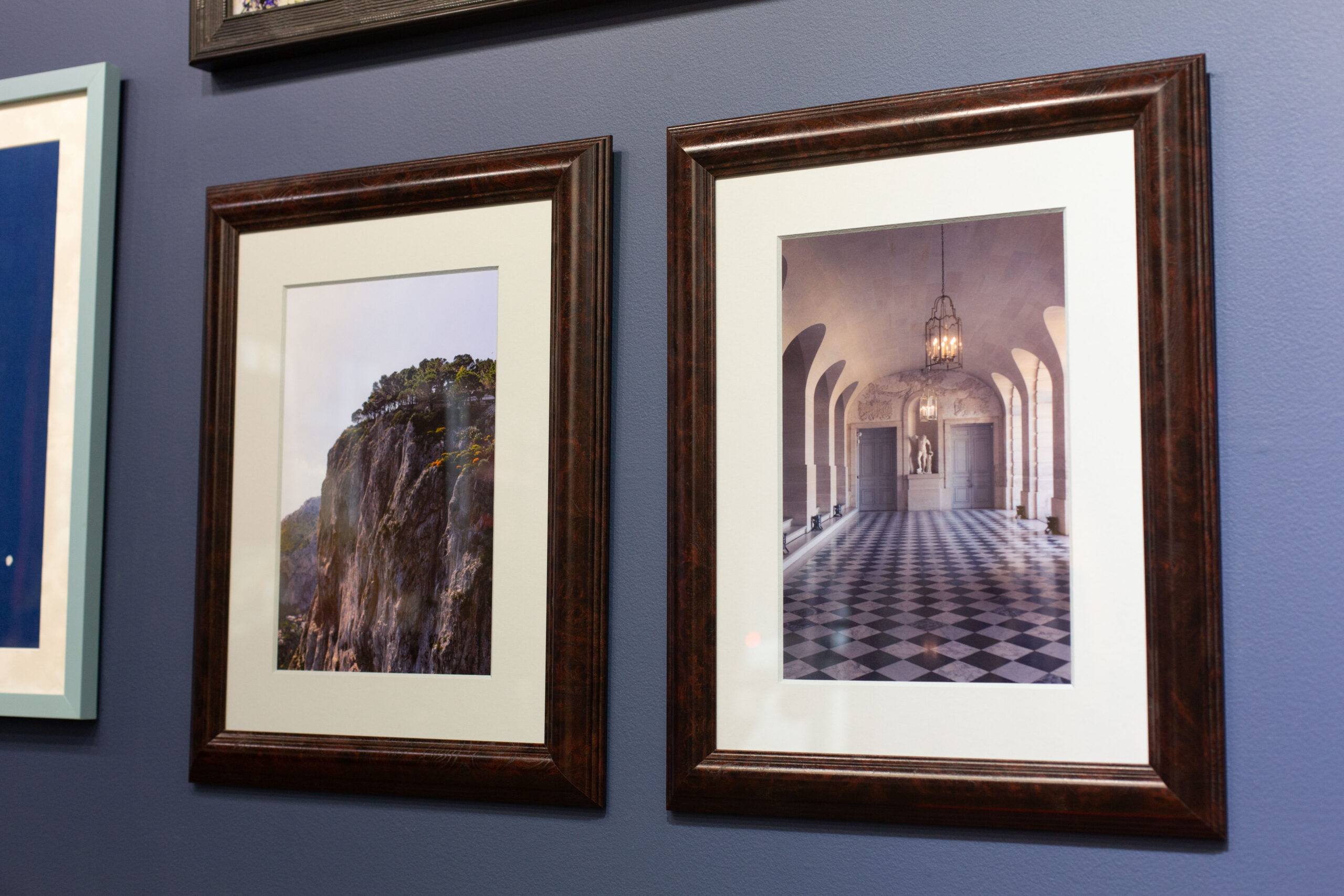 Two photographs framed in rich-colored wood frames hang on the gallery wall. One photograph is a print of a cliffside, while the other is an elegant hallway.