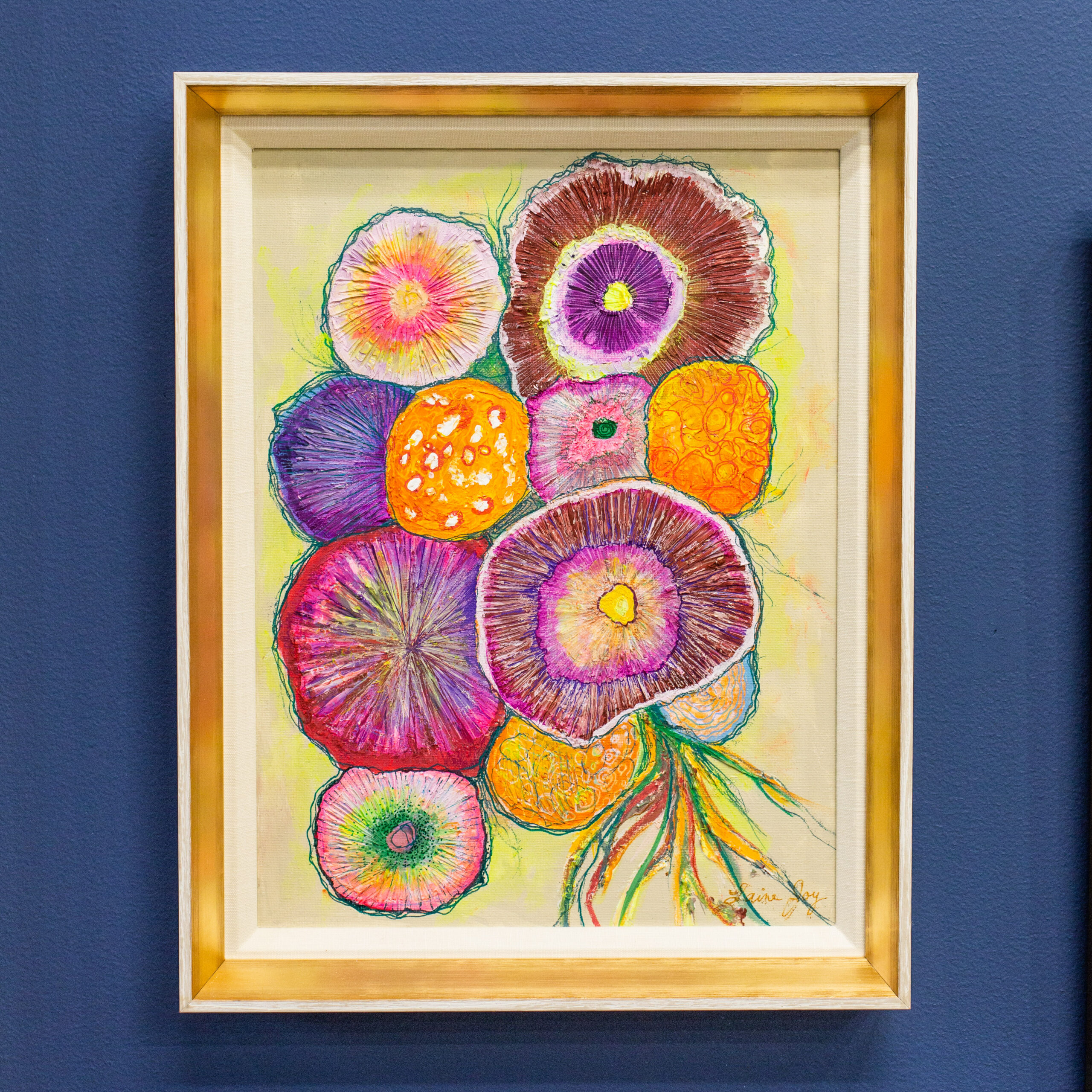 A closeup of a beautiful, colorful painting of a bouquet framed in a white and light brown frame. The painting hangs from a blue gallery wall.