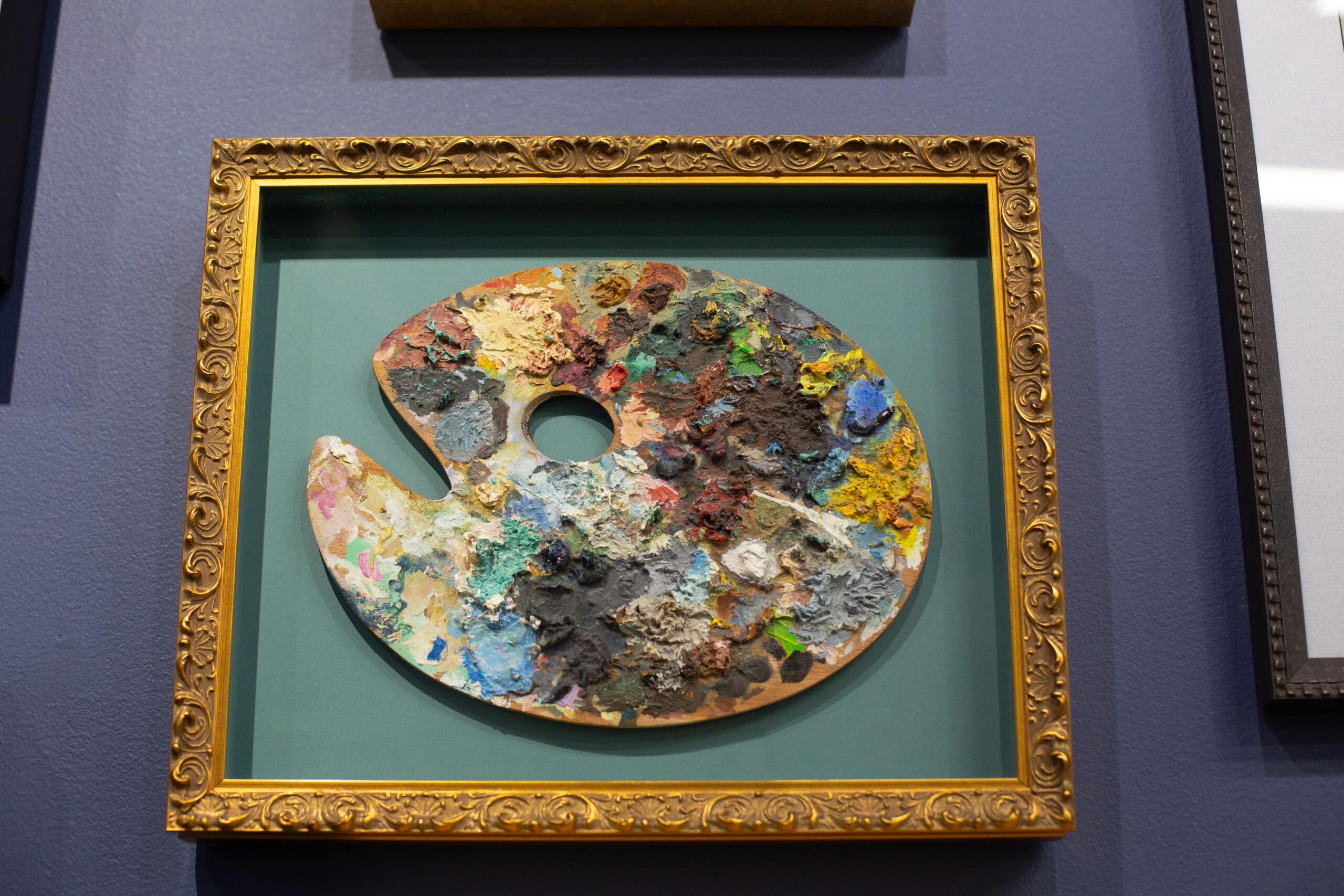 A closeup of an artist's paint palette covered in paint and framed in an elegant gold frame. The painting hangs from a blue gallery wall.