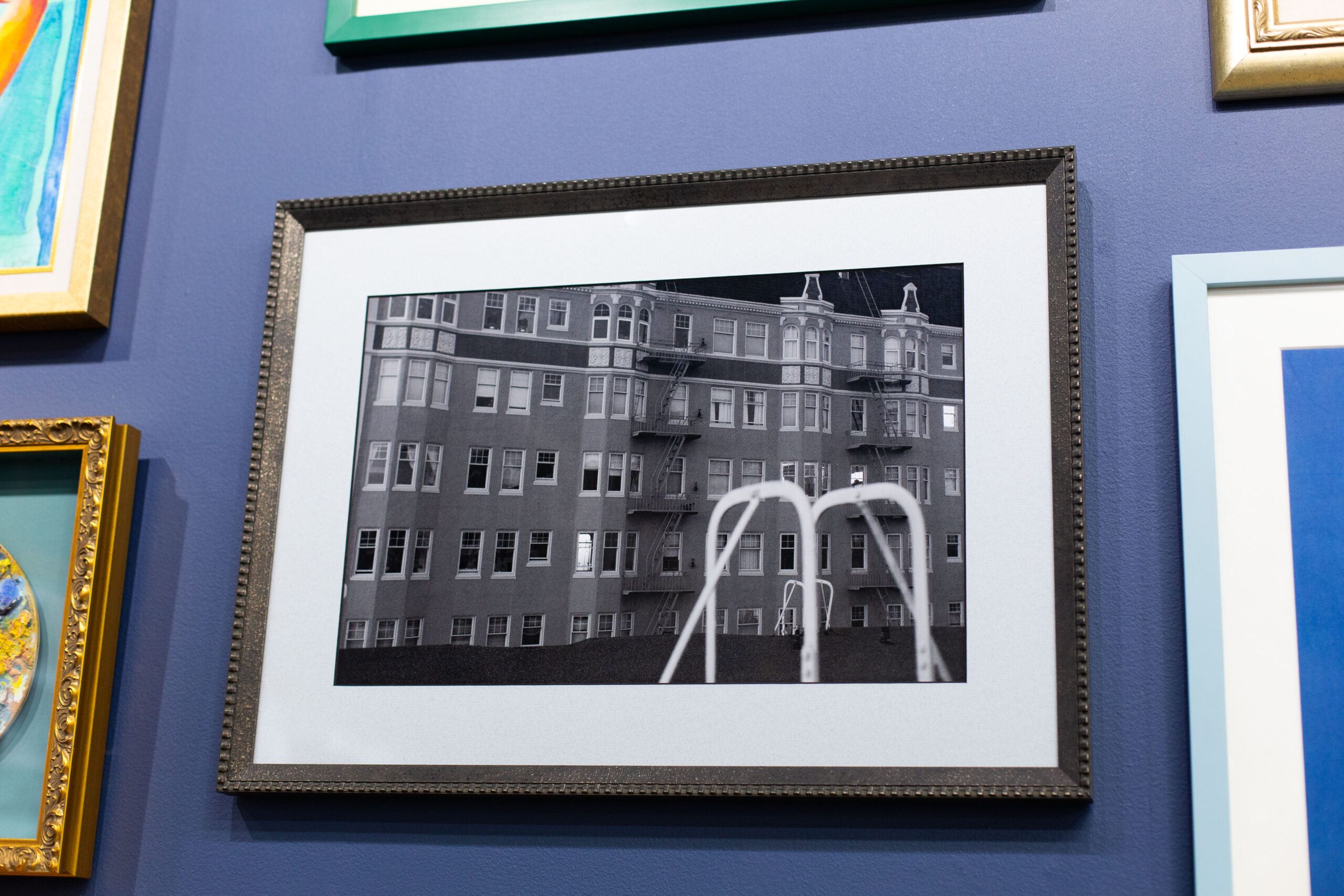 A closeup of a black and white photograph of the outside of an apartment building. The photo hangs from a blue wall in the Peterson Picture Co. gallery.