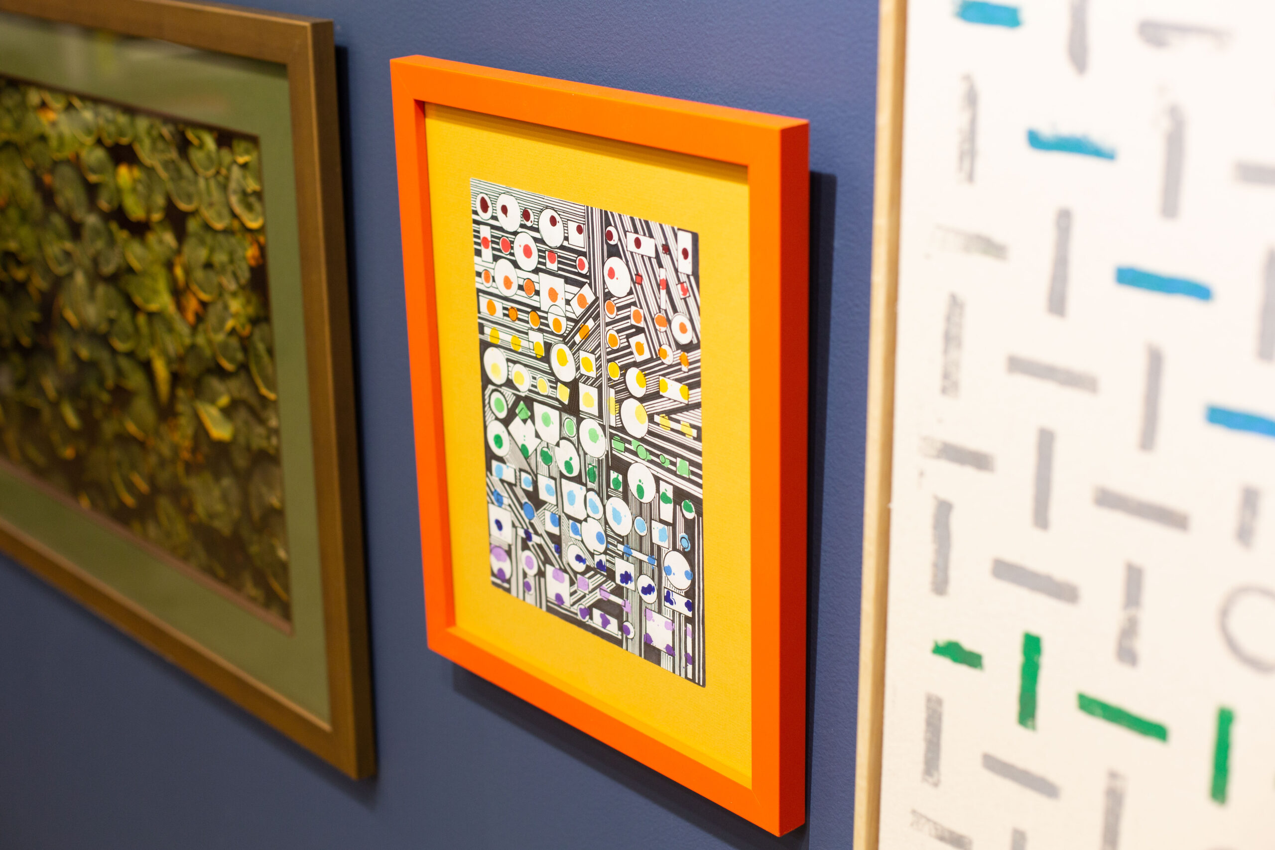 A closeup angle shows three pieces of art, focusing on the drawing in the middle in a bright orange frame. The photos hang in the Peterson Picture Co. gallery.