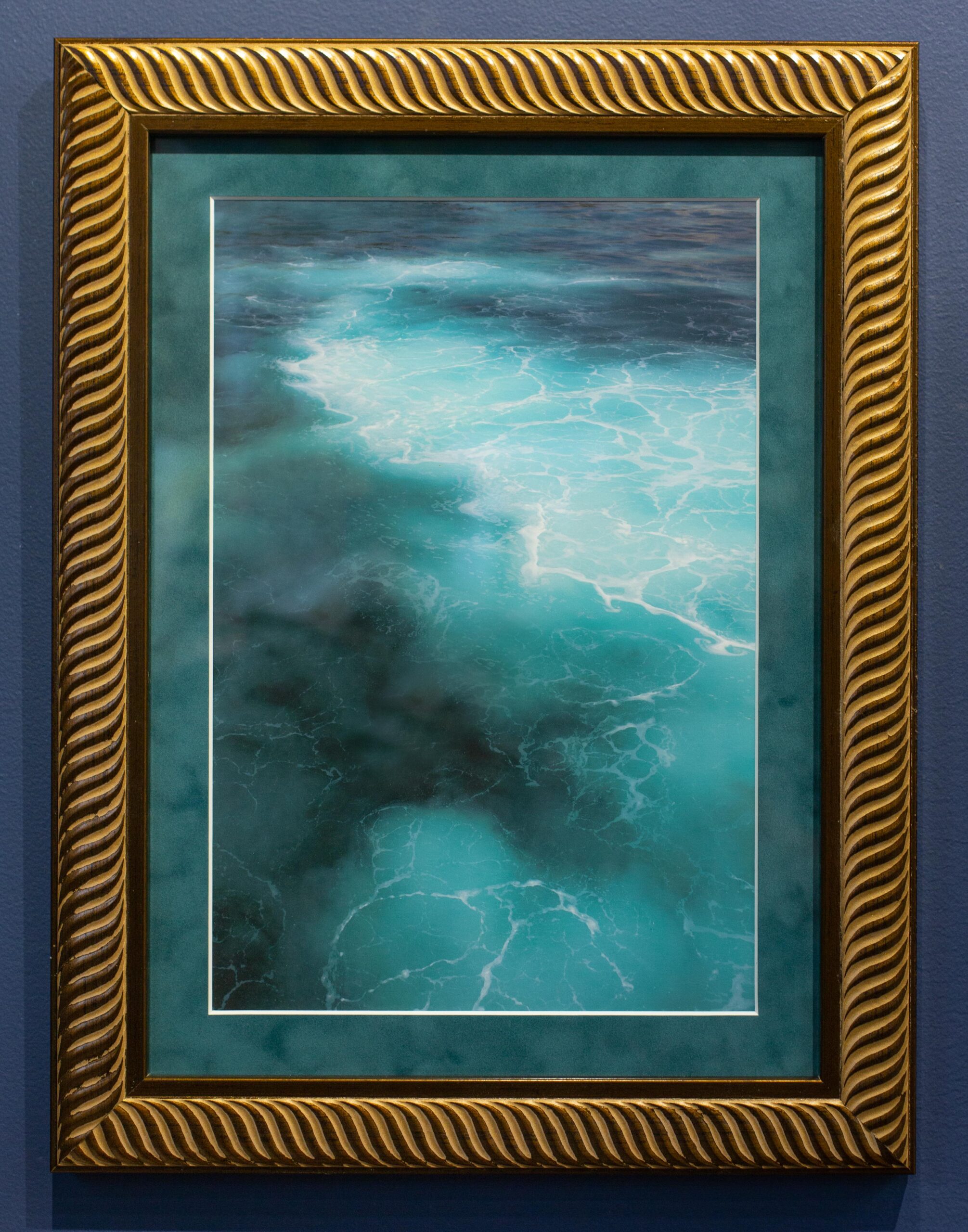 A closeup shows an image of a moody ocean from an aerial viewpoint. The art is in a gold frame and is mounted on a wall in the Peterson Picture Co. gallery.